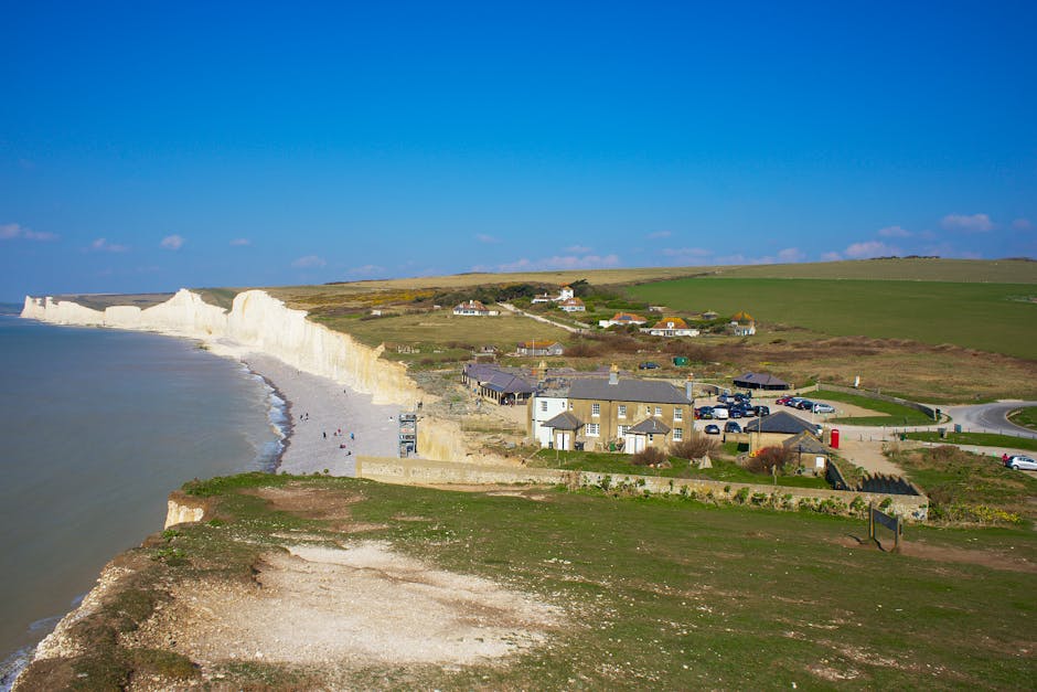 A coastal scene featuring white chalk cliffs towering above a small seaside village with a mix of residential buildings, some with pitched roofs and light-colored facades. The village is situated on g