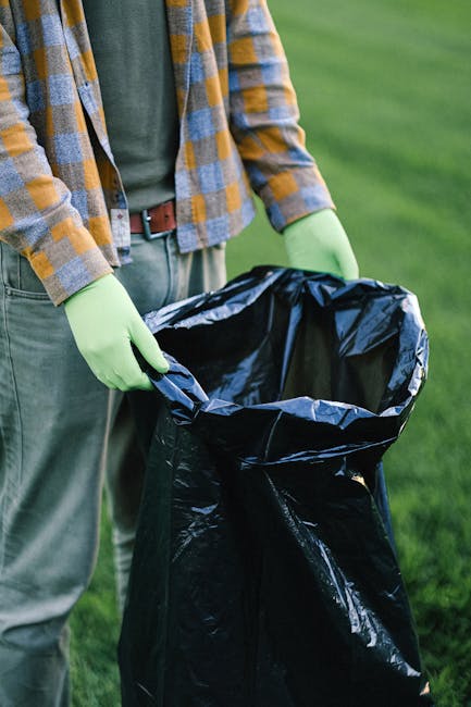 A sanitation worker wearing a high-visibility vest is unloading waste from the rear compartments of a large red garbage truck parked beside a street. The truck, identified by its mechanical components